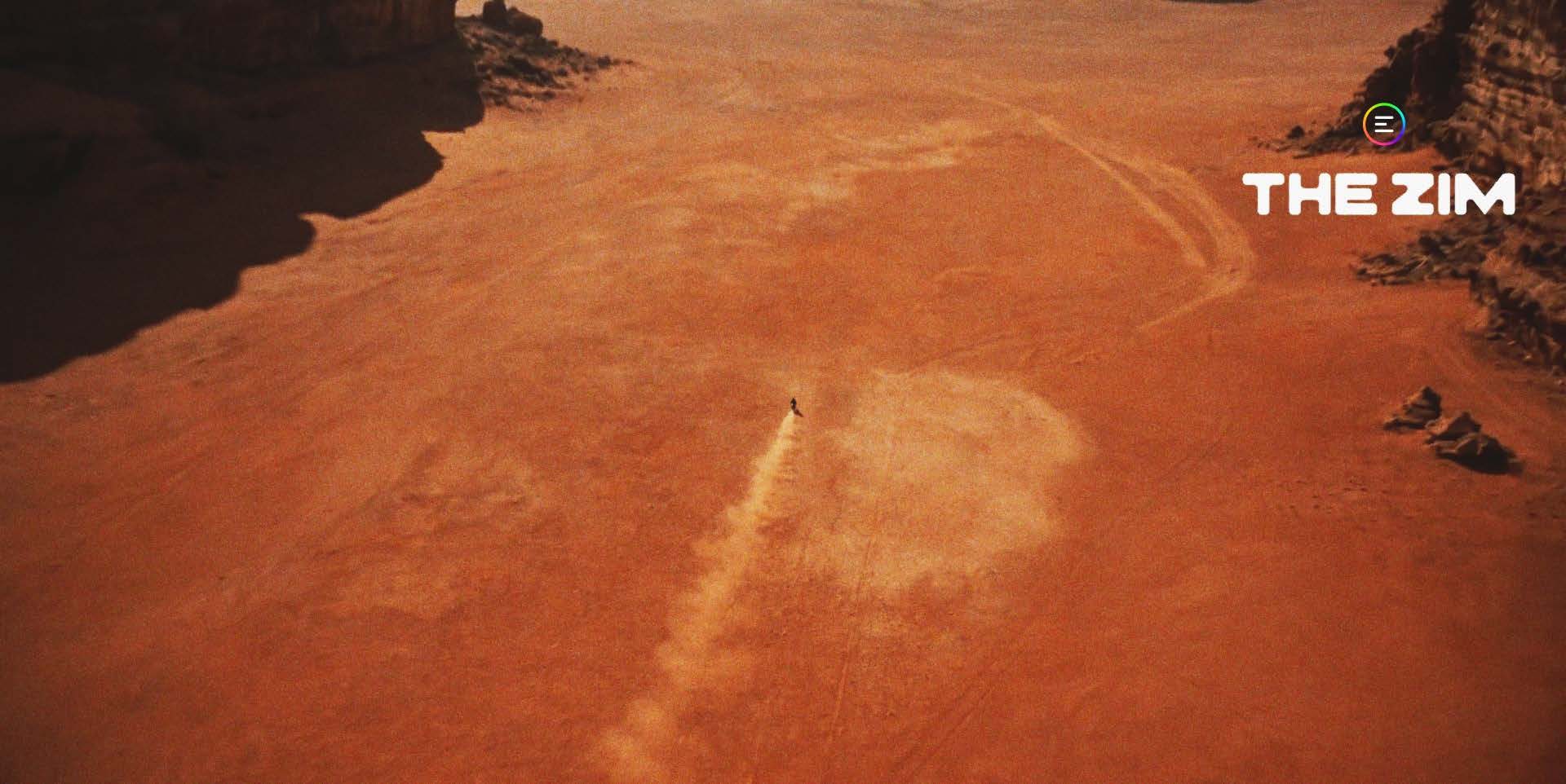 High aerial shot of a vast red-orange desert basin. A single tiny dark figure or craft crosses the open sand near the centre of frame, kicking up a long pale dust plume that trails down and to the left behind it. A broad